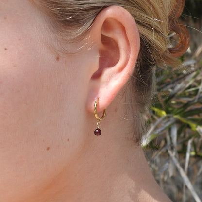 A close-up of a person's ear wearing a small gold hoop earring with a dangling dark red gemstone, set against an outdoor background with blurred foliage.