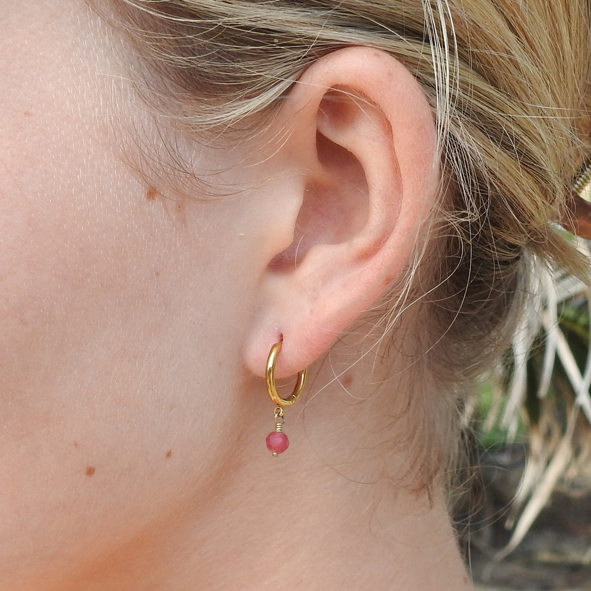 A close-up of a person's ear wearing a small gold hoop earring with a dangling pink bead.