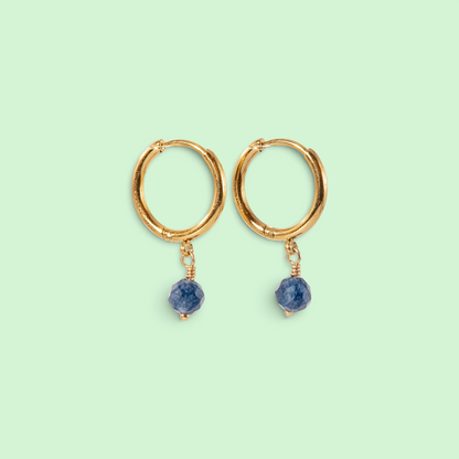 A pair of gold huggie hoop earrings with small faceted blue gemstone dangles, shown on a solid pale mint green background.