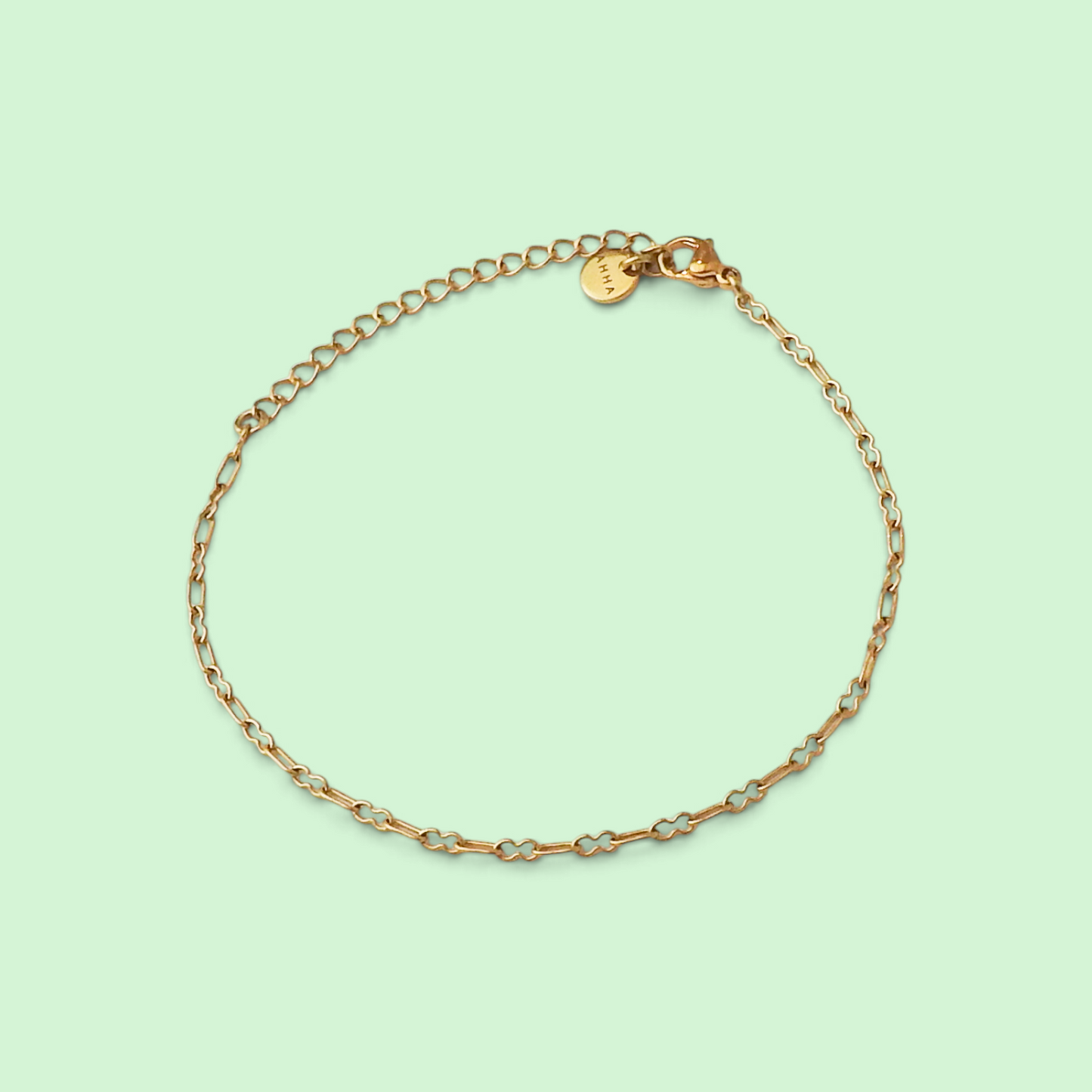 A delicate gold chain bracelet with alternating small gold links and thin silver-toned wire accents, displayed on a solid pale mint green background. A small round gold tag is attached to the clasp.