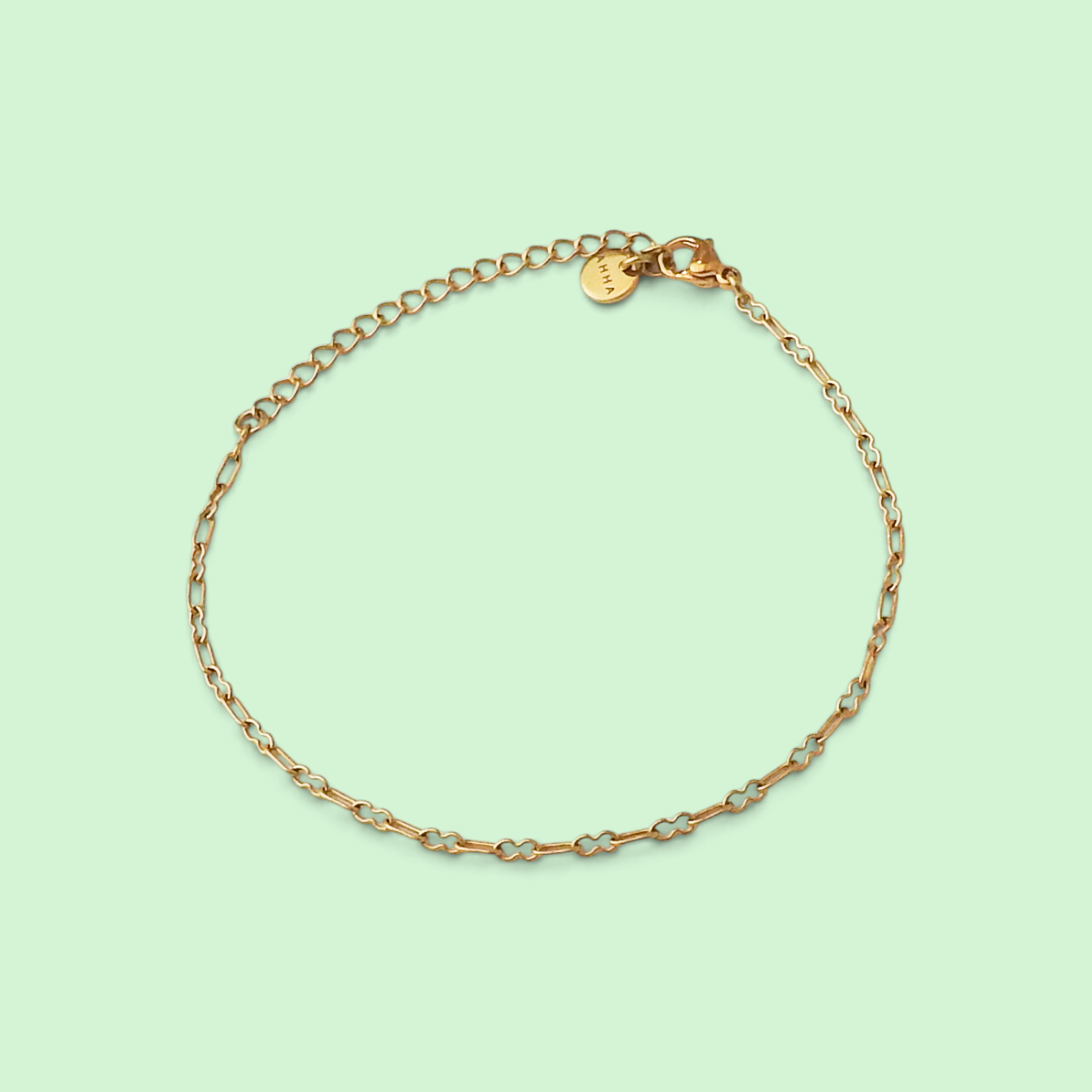 A delicate gold chain bracelet with alternating small gold links and thin silver-toned wire accents, displayed on a solid pale mint green background. A small round gold tag is attached to the clasp.