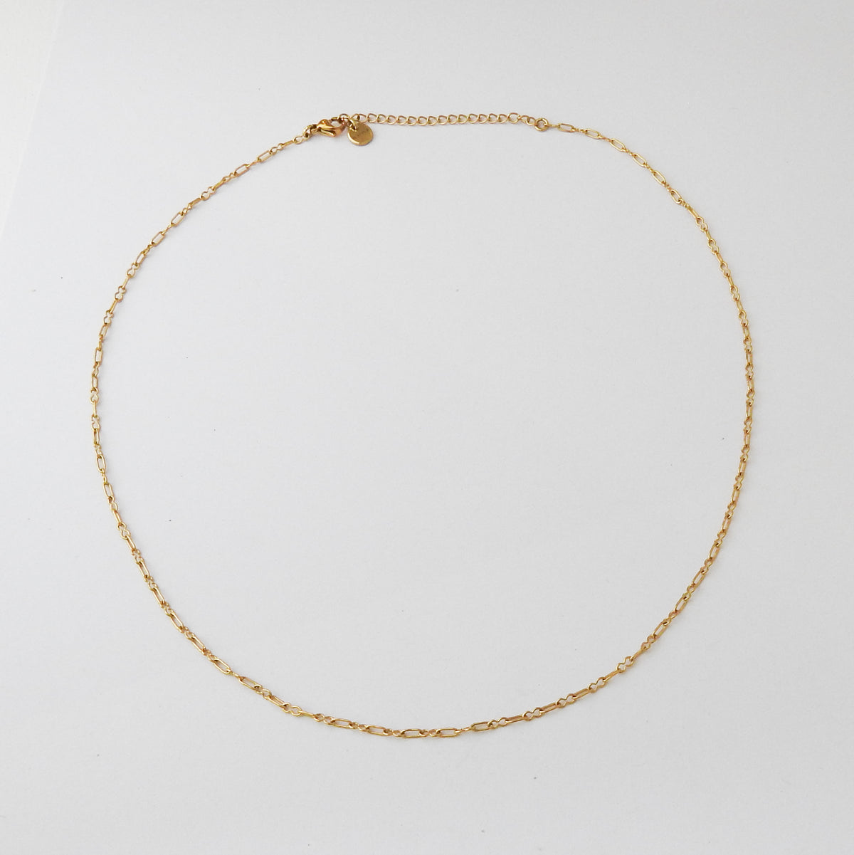 A delicate gold chain necklace with a small oval charm, arranged in a circle on a plain white background.