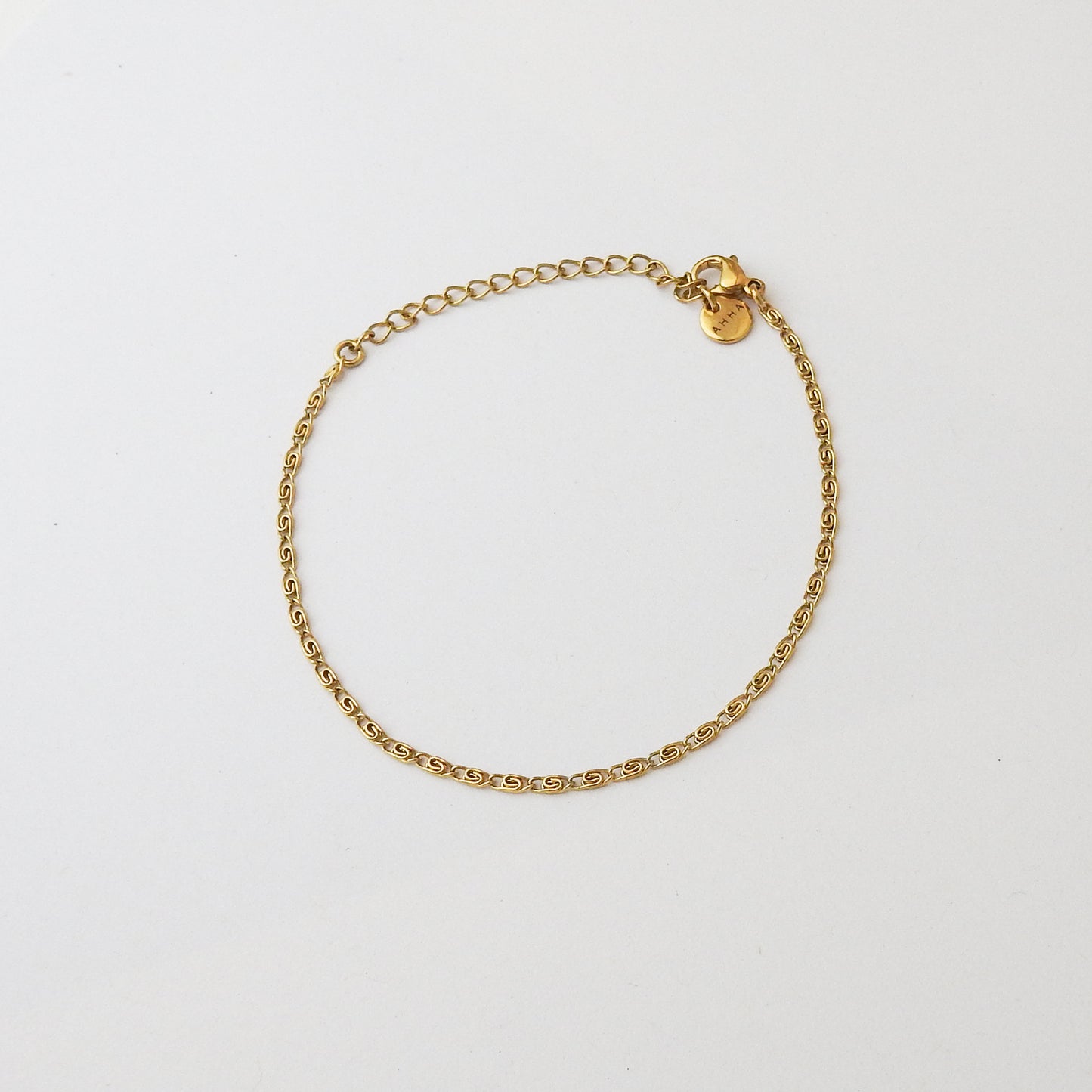 A delicate gold chain bracelet with a small lobster clasp and a round gold tag, displayed on a clean white background.