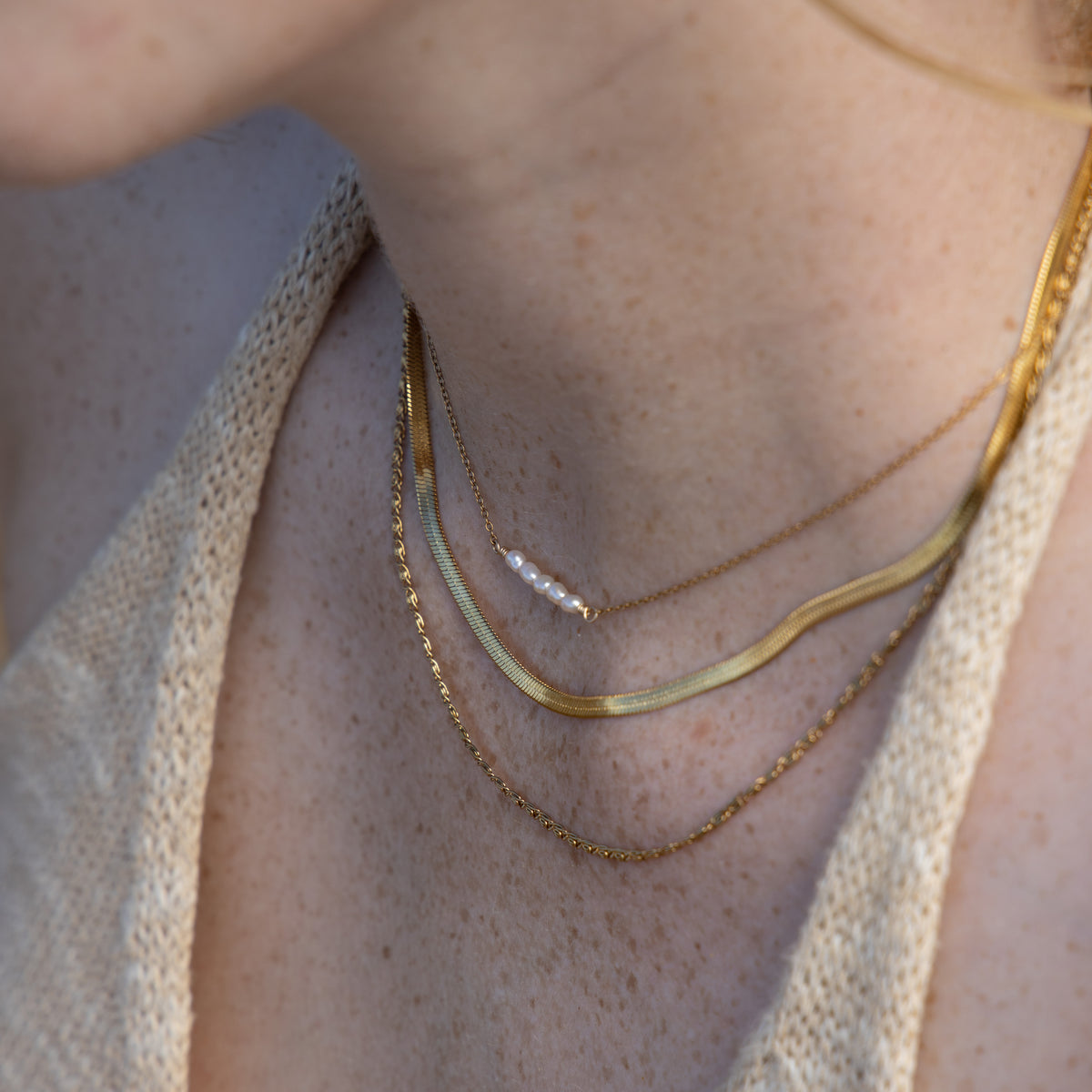 Close-up of a person's neck and upper chest wearing layered gold necklaces, including a thin chain with a small pearl accent, on a beige knit top.