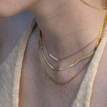 Close-up of a person's neck and upper chest wearing layered gold necklaces, including a thin chain with a small pearl accent, on a beige knit top.