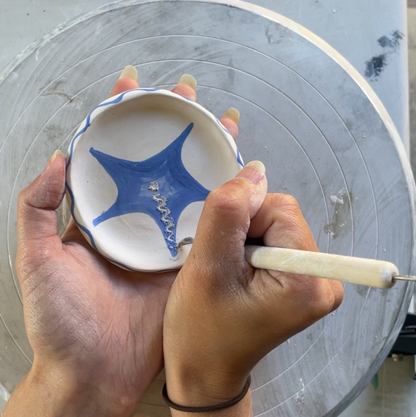 A close-up of a person's hands holding a small white ceramic bowl with a blue star design while using a pottery tool on the inside of the bowl, set against a pottery wheel background.