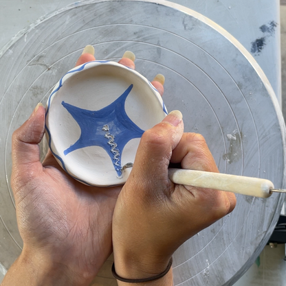 A close-up of a person's hands holding a small white ceramic bowl with a blue star design while using a pottery tool on the inside of the bowl, set against a pottery wheel background.