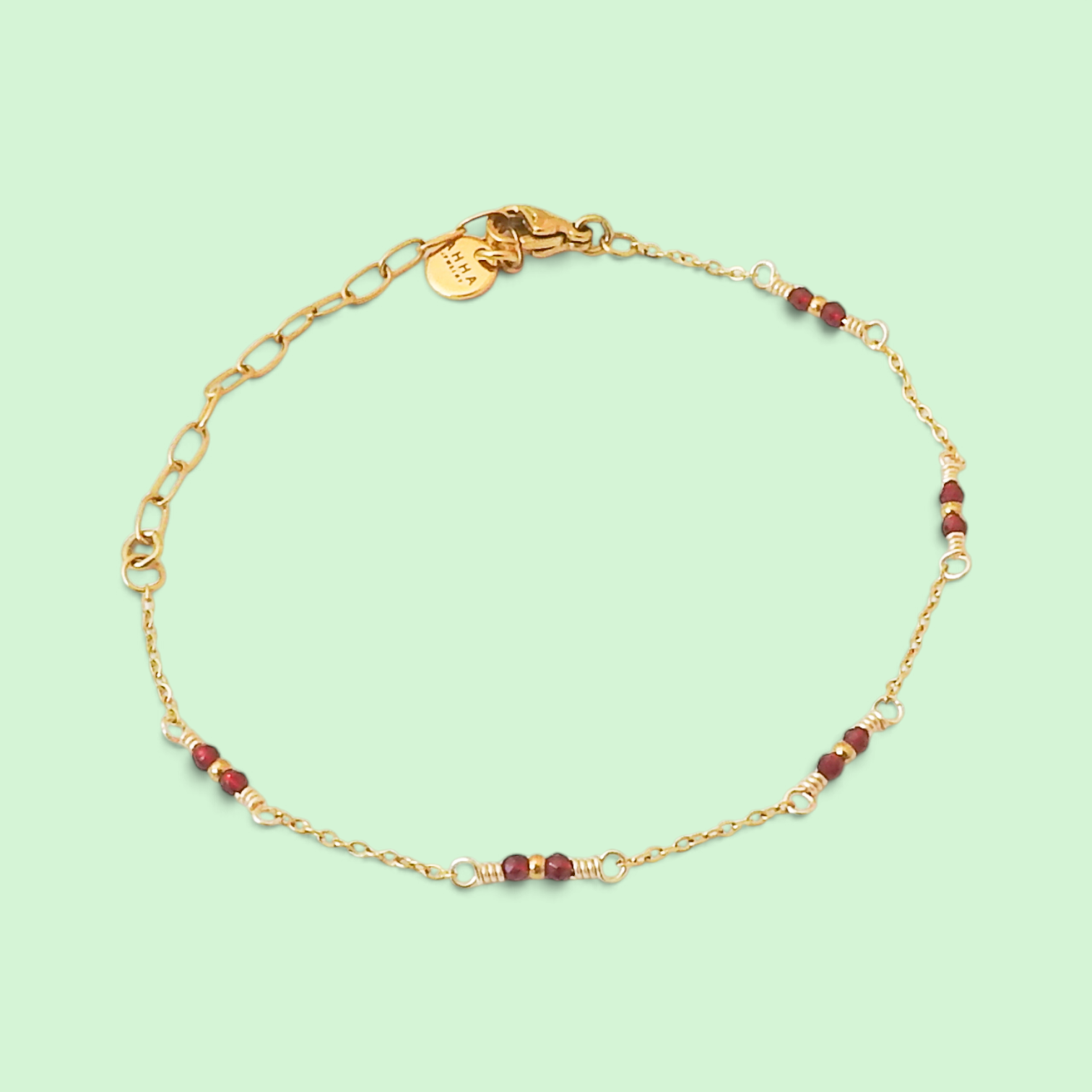 A delicate gold chain bracelet with small red beads and silver-toned spacer accents, displayed on a light mint green background.