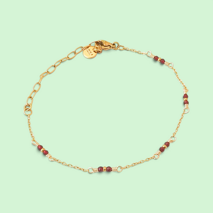 A delicate gold chain bracelet with small red beads and silver-toned spacer accents, displayed on a light mint green background.