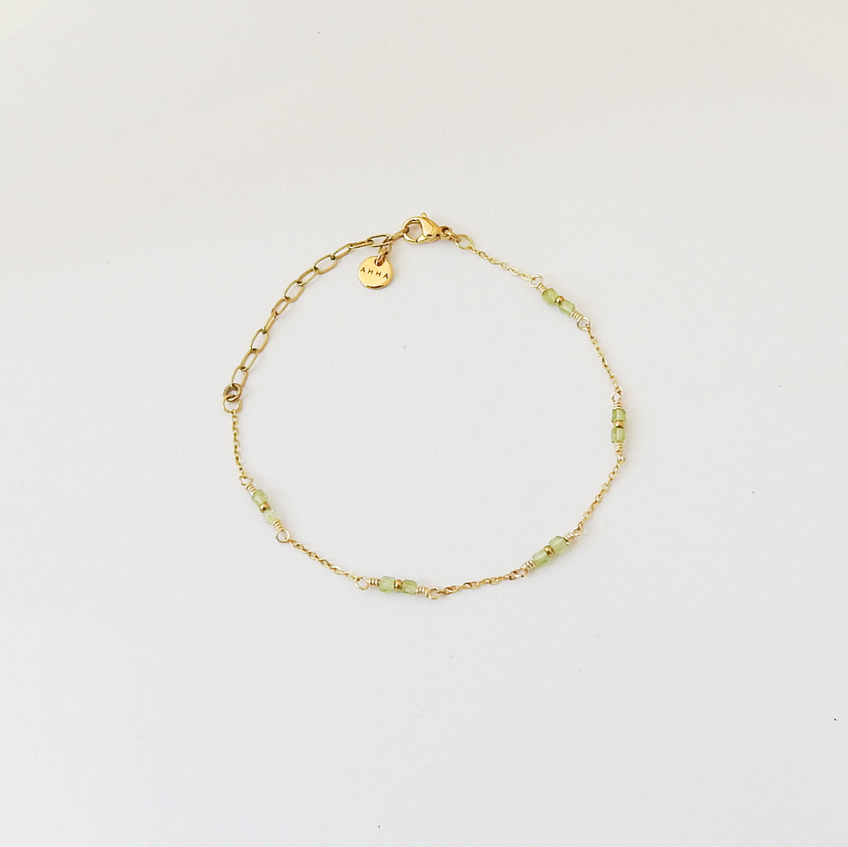 A delicate gold chain bracelet with small light green beads and a small heart-shaped charm, displayed on a clean white background.