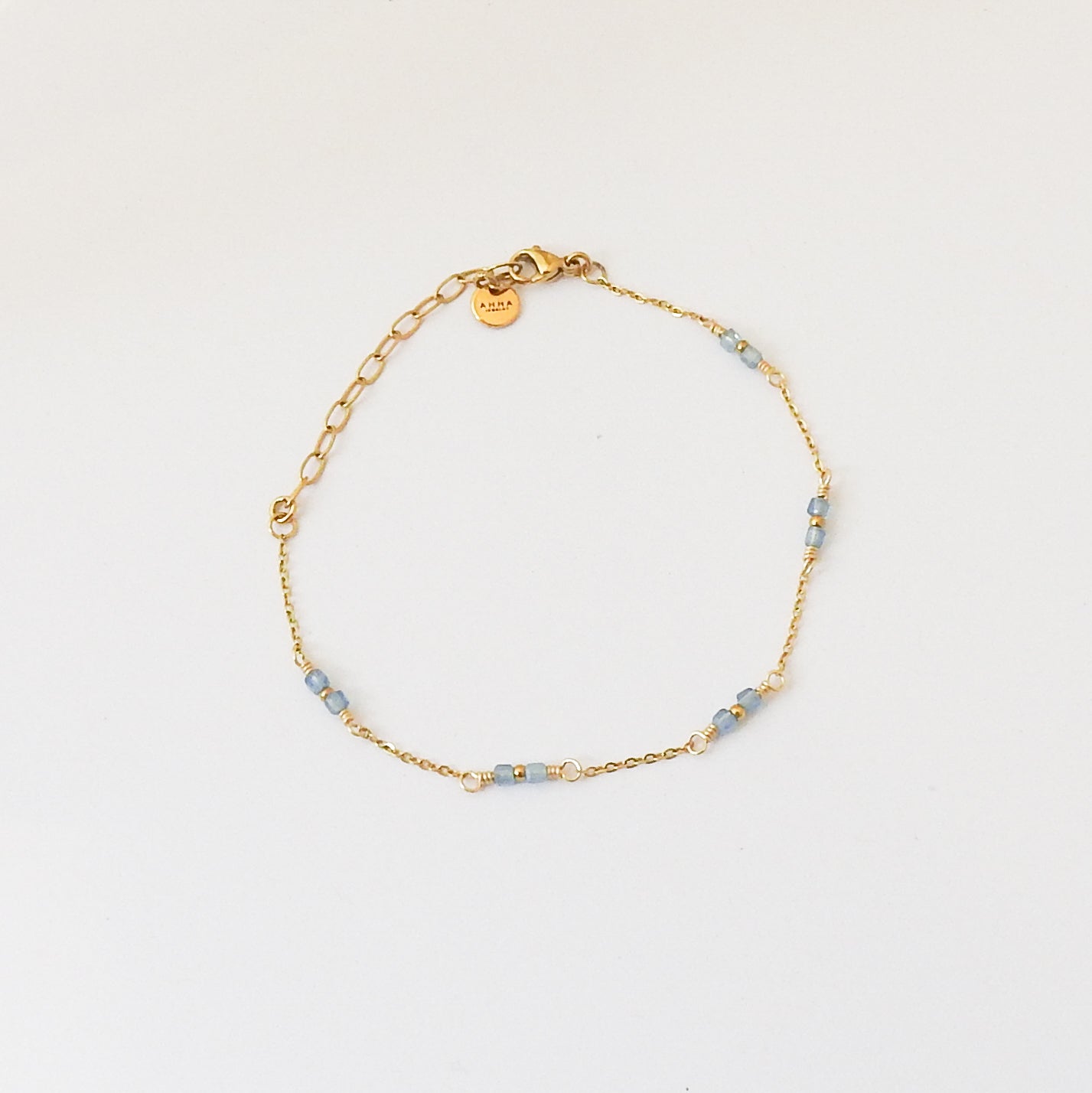 A delicate gold chain bracelet with small light blue beads and a small round gold tag, displayed on a clean white background.