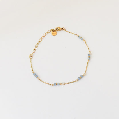 A delicate gold chain bracelet with small light blue beads and a small round gold tag, displayed on a clean white background.