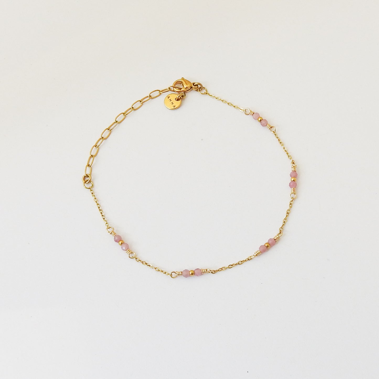 A delicate gold chain bracelet with small pink faceted beads spaced along the front, displayed on a clean white background. It features an adjustable lobster clasp and a small round gold tag.