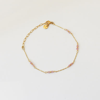 A delicate gold chain bracelet with small pink faceted beads spaced along the front, displayed on a clean white background. It features an adjustable lobster clasp and a small round gold tag.
