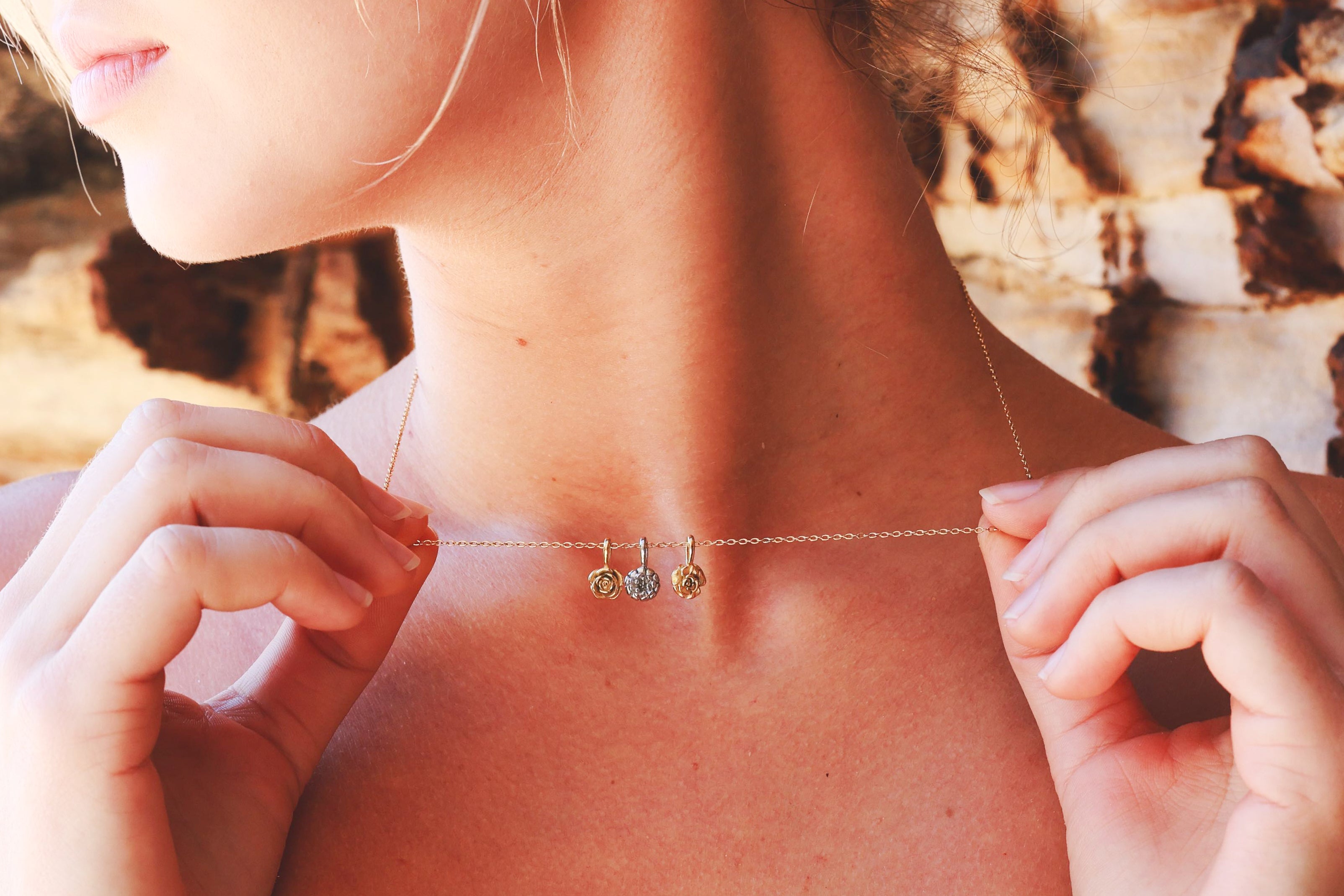 A close-up lifestyle shot of a model wearing a delicate gold chain necklace with three small dangling charms, including a clear crystal and two gold beads, against a rustic wood background.