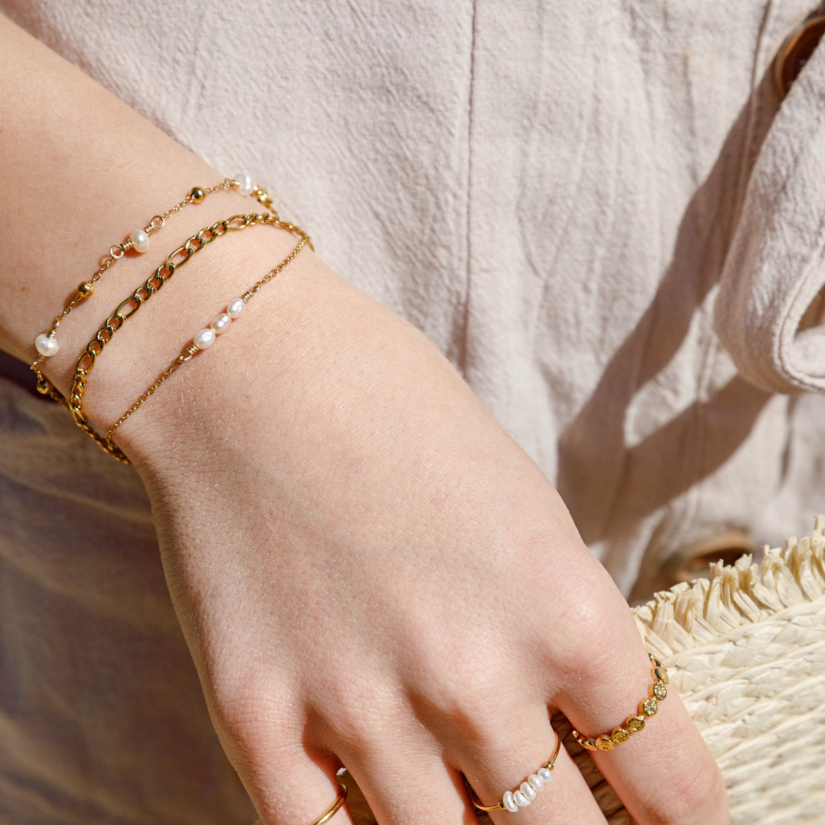 A close-up of a person's hand wearing delicate gold jewelry, including a double-strand gold chain bracelet with small white pearls, a thin gold chain bracelet, and several gold rings, resting on a straw hat.