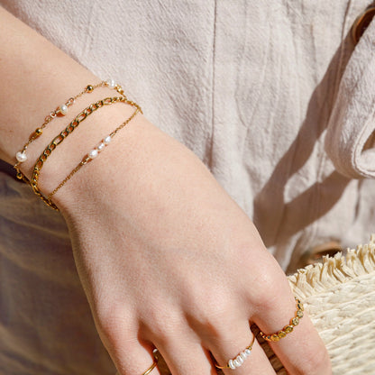 A close-up of a person's hand wearing delicate gold jewelry, including a double-strand gold chain bracelet with small white pearls, a thin gold chain bracelet, and several gold rings, resting on a straw hat.