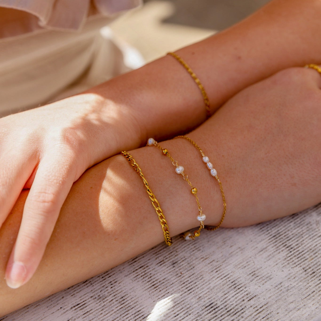 A close-up of a person's forearm adorned with three delicate gold bracelets, including a curb chain, a beaded chain with white pearls, and a thin chain with small gold beads.