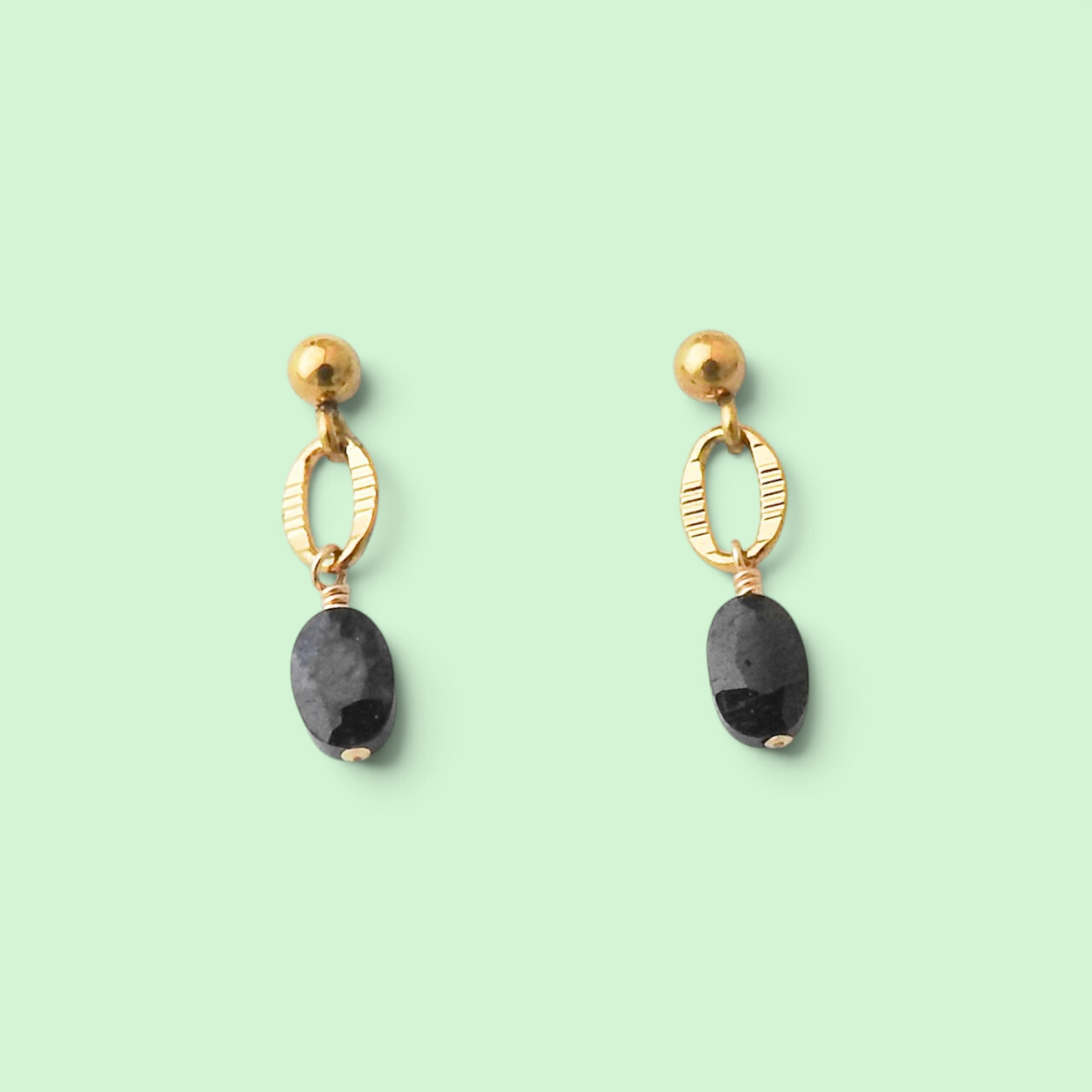 A pair of gold drop earrings with textured oval hoops, gold ball studs, and dark gray faceted oval gemstones, displayed on a solid pale green background.