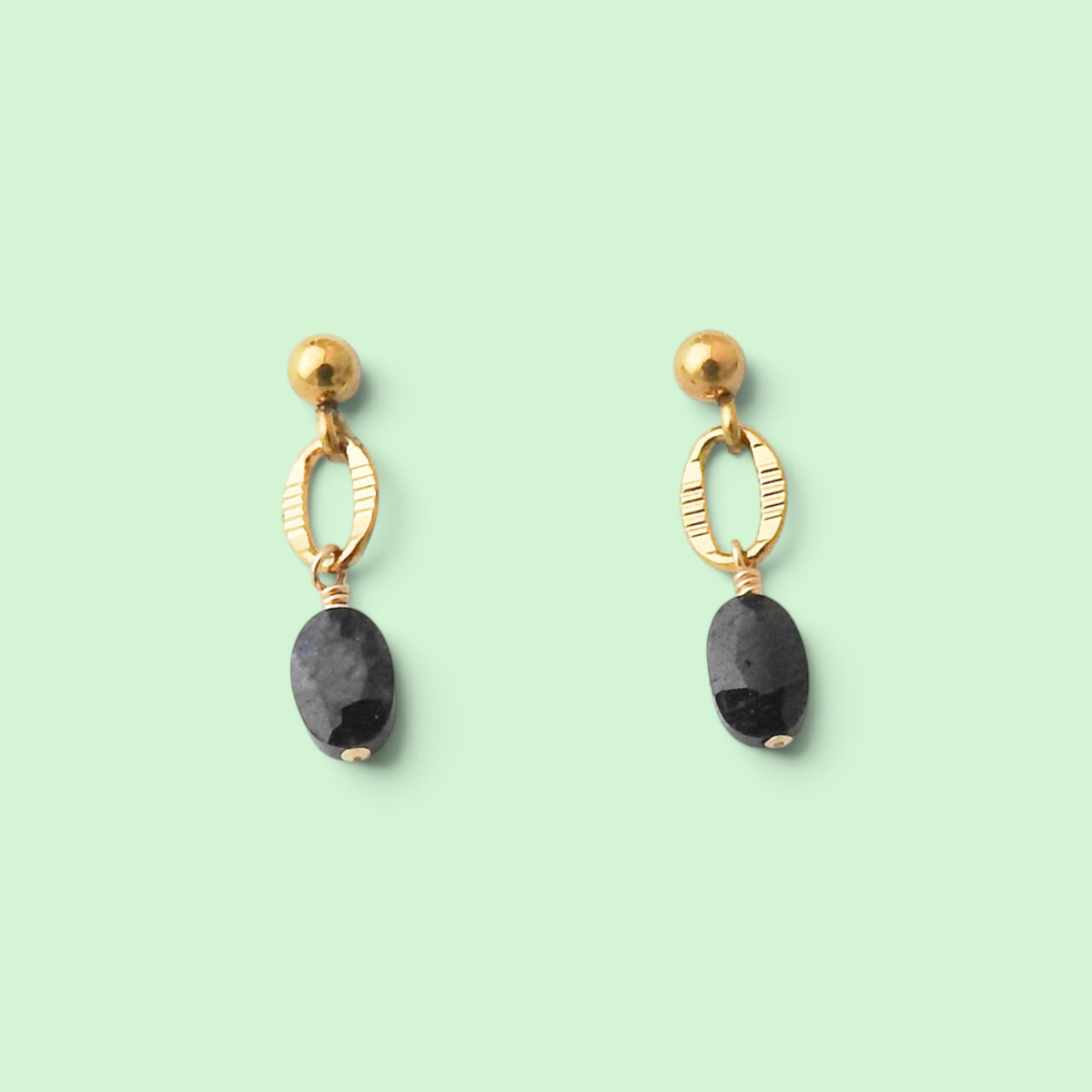 A pair of gold drop earrings with textured oval hoops, gold ball studs, and dark gray faceted oval gemstones, displayed on a solid pale green background.