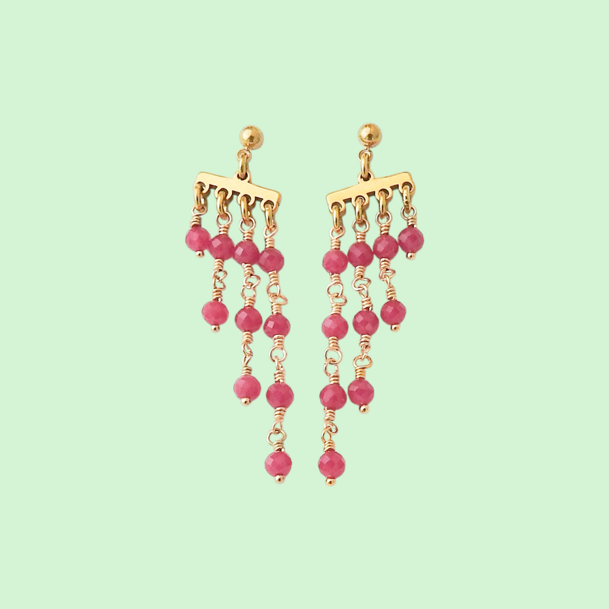 A pair of gold-tone dangle earrings with multiple cascading chains of pink faceted beads, shown against a solid pale mint green background.