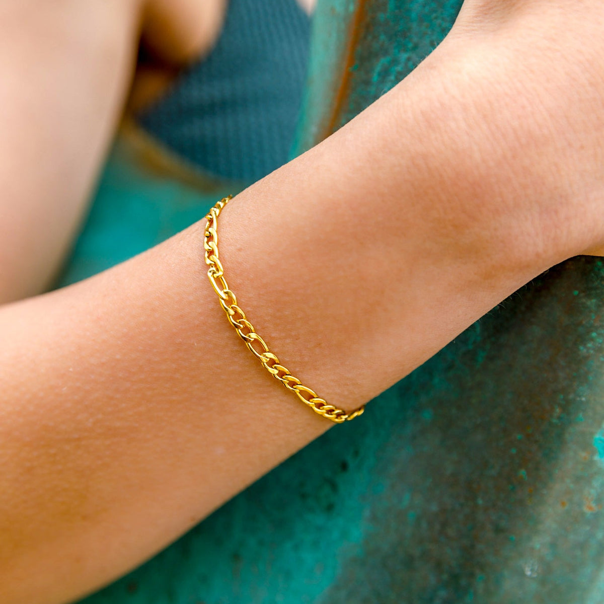 A close-up of a delicate gold chain bracelet worn on a person's wrist, resting against a textured teal metal surface.