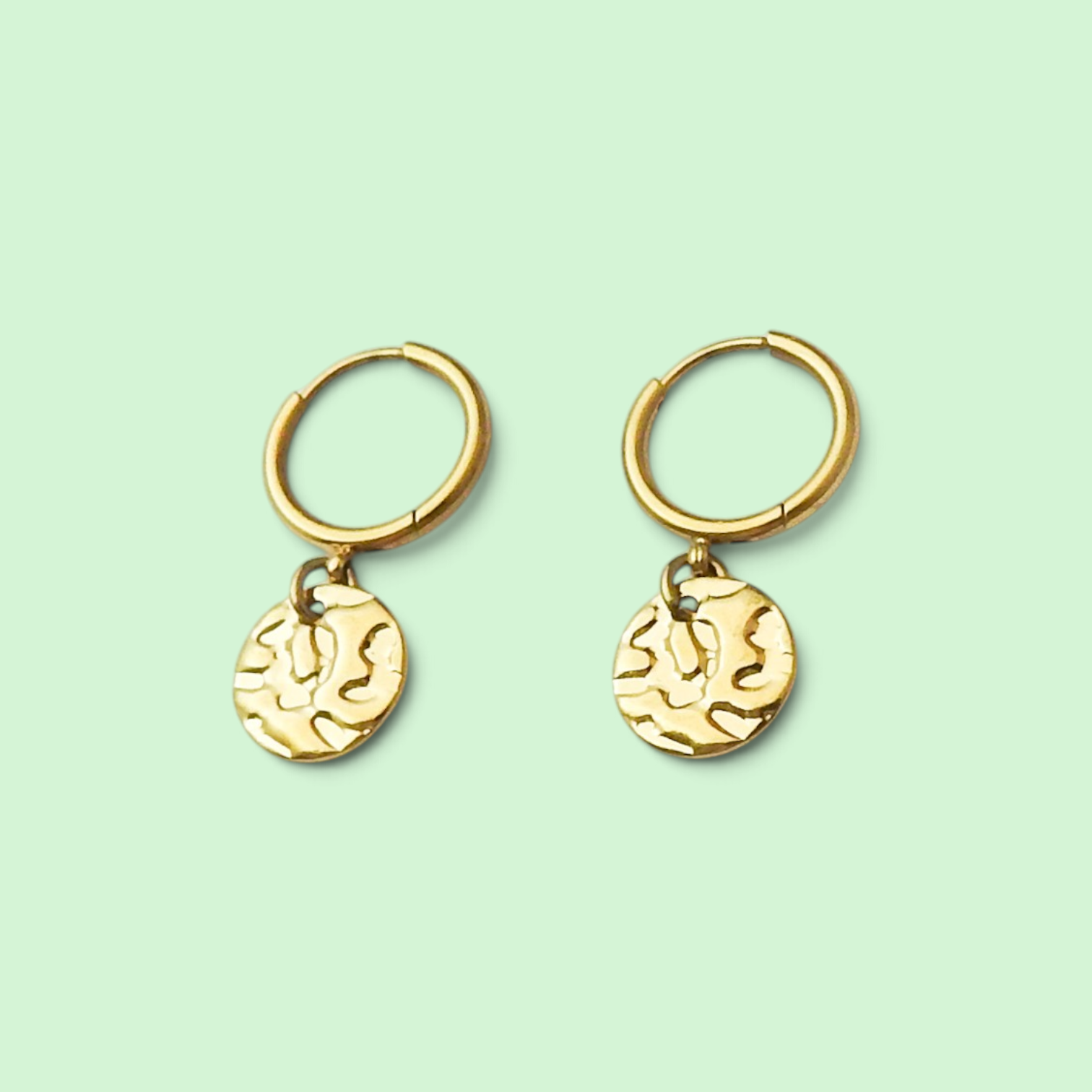 A pair of gold hoop earrings with textured, organic-shaped round dangles, displayed on a solid pale mint green background.