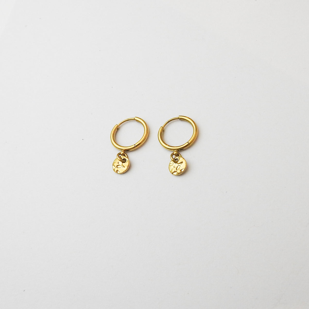 A pair of small gold huggie hoop earrings with dangling textured circular charms, displayed on a clean white background.