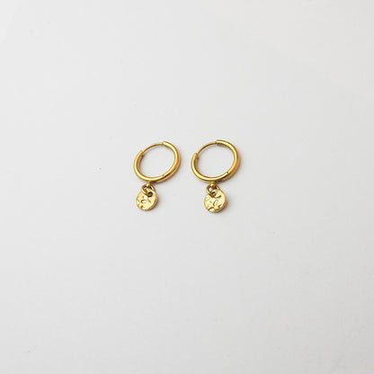 A pair of small gold huggie hoop earrings with dangling textured circular charms, displayed on a clean white background.