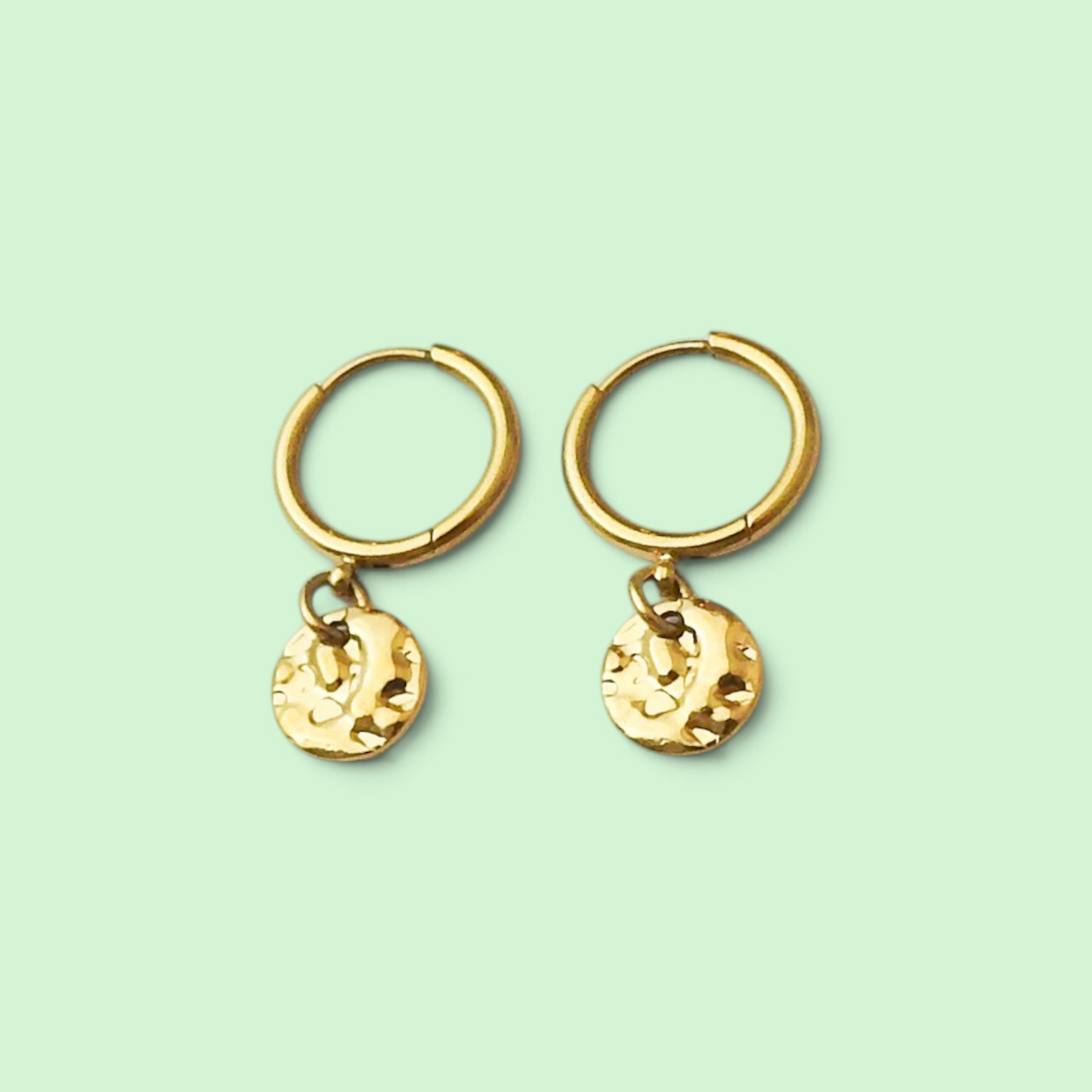 A pair of gold huggie hoop earrings with textured round disc charms, displayed on a solid pale mint green background.