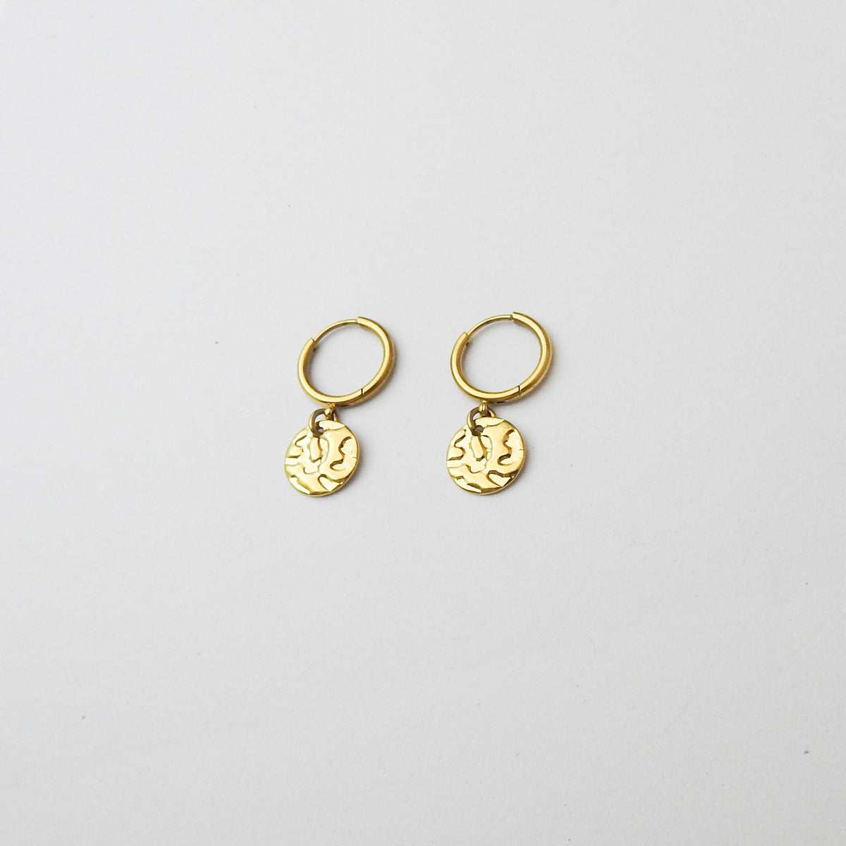 A pair of small gold huggie hoop earrings with dangling textured round charms, displayed on a clean white background.