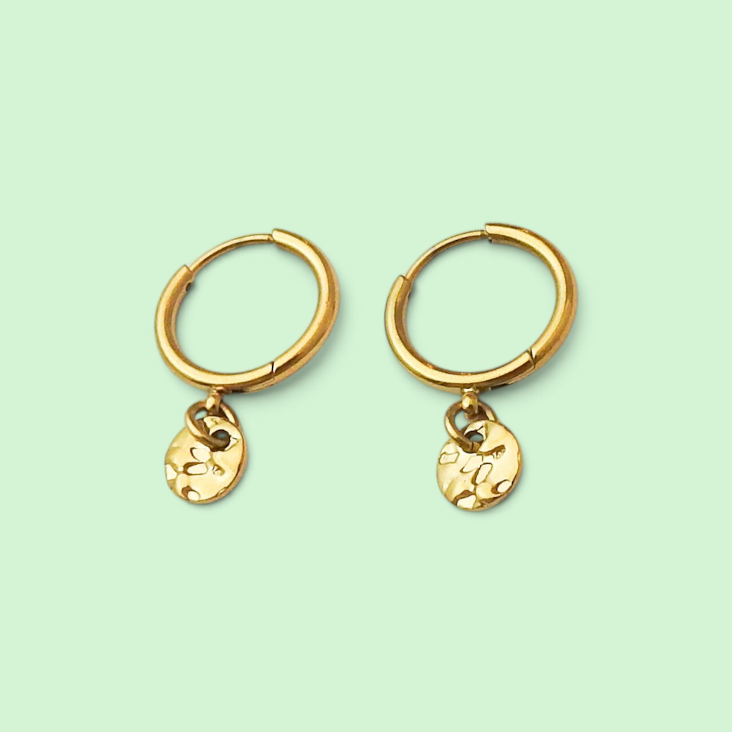 A pair of gold hoop earrings with small, textured oval dangling charms, displayed on a solid pale mint green background.