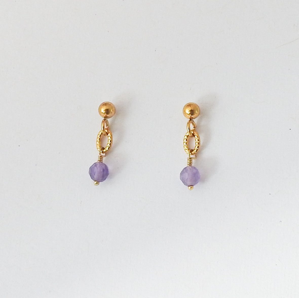 A pair of gold dangle earrings with a faceted light purple bead at the bottom, displayed on a clean white background.