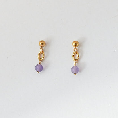 A pair of gold dangle earrings with a faceted light purple bead at the bottom, displayed on a clean white background.