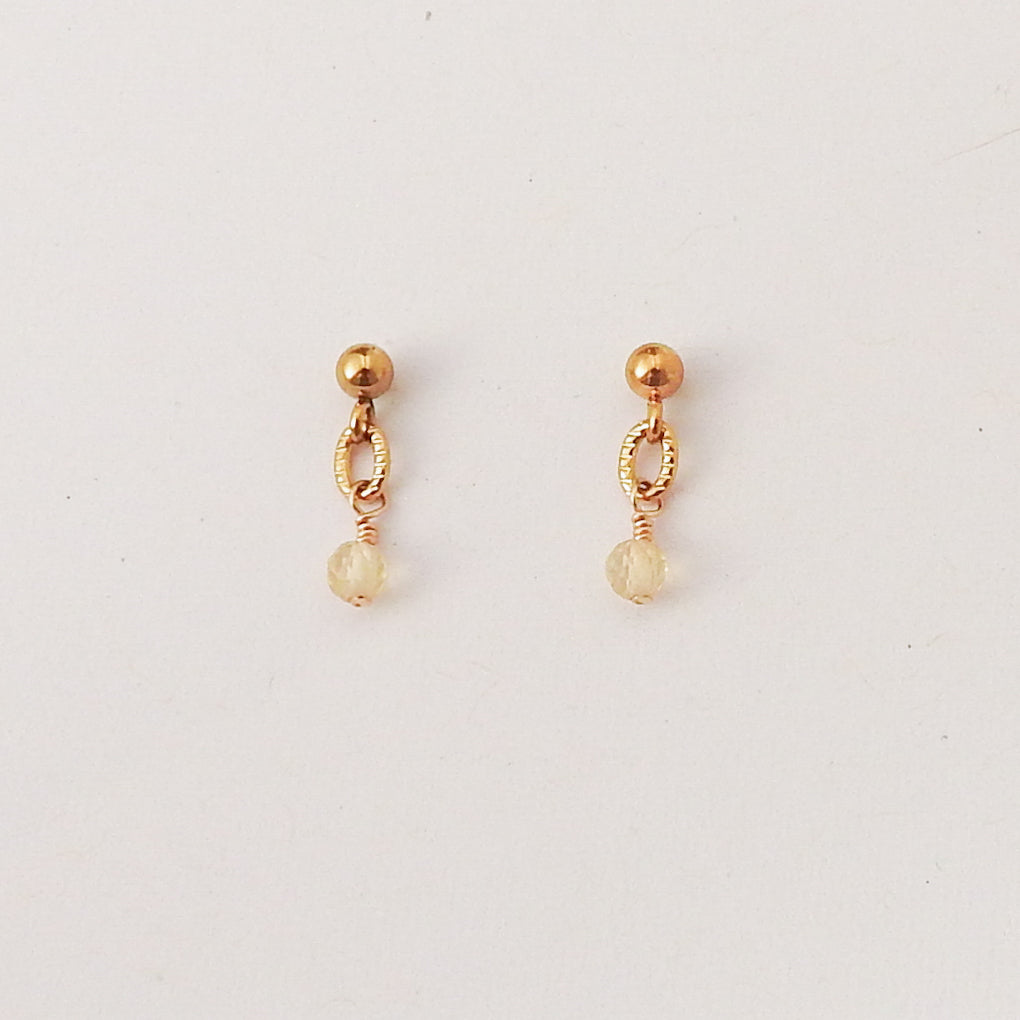 A pair of gold-toned dangle earrings with a small round bead, textured oval link, and faceted clear crystal drop, shown on a plain light background.