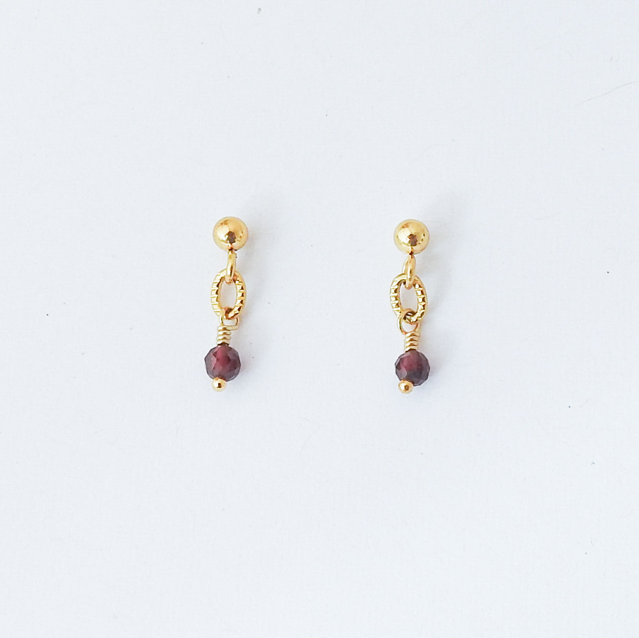 A pair of gold dangle earrings with a small gold ball stud, a textured oval link, a thin wire wrap, and a faceted dark red bead drop, shown on a plain white background.