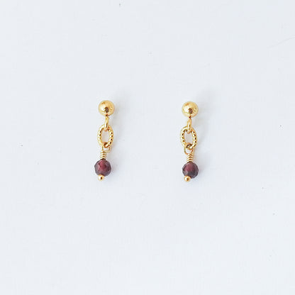 A pair of gold dangle earrings with a small gold ball stud, a textured oval link, a thin wire wrap, and a faceted dark red bead drop, shown on a plain white background.