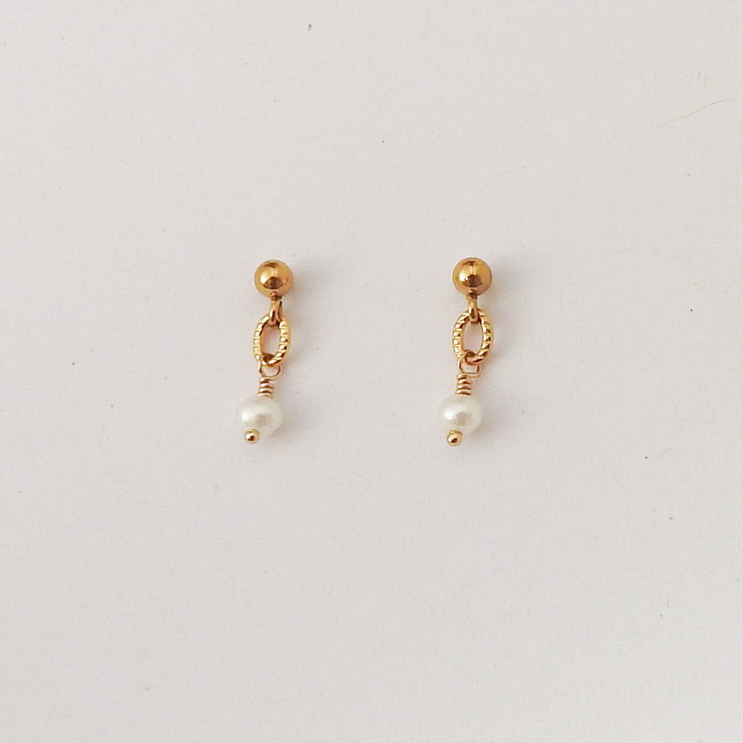 A pair of gold dangle earrings with a small gold bead, twisted oval loop, and a dangling white pearl on a plain light background.