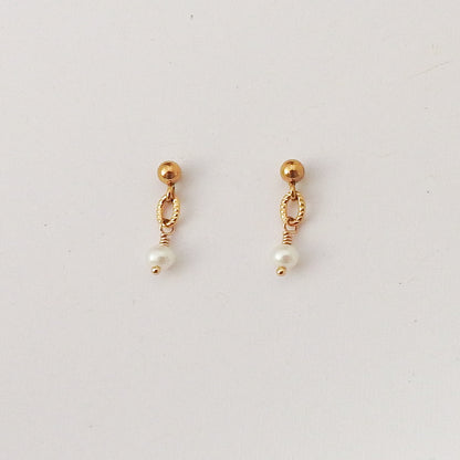 A pair of gold dangle earrings with a small gold bead, twisted oval loop, and a dangling white pearl on a plain light background.