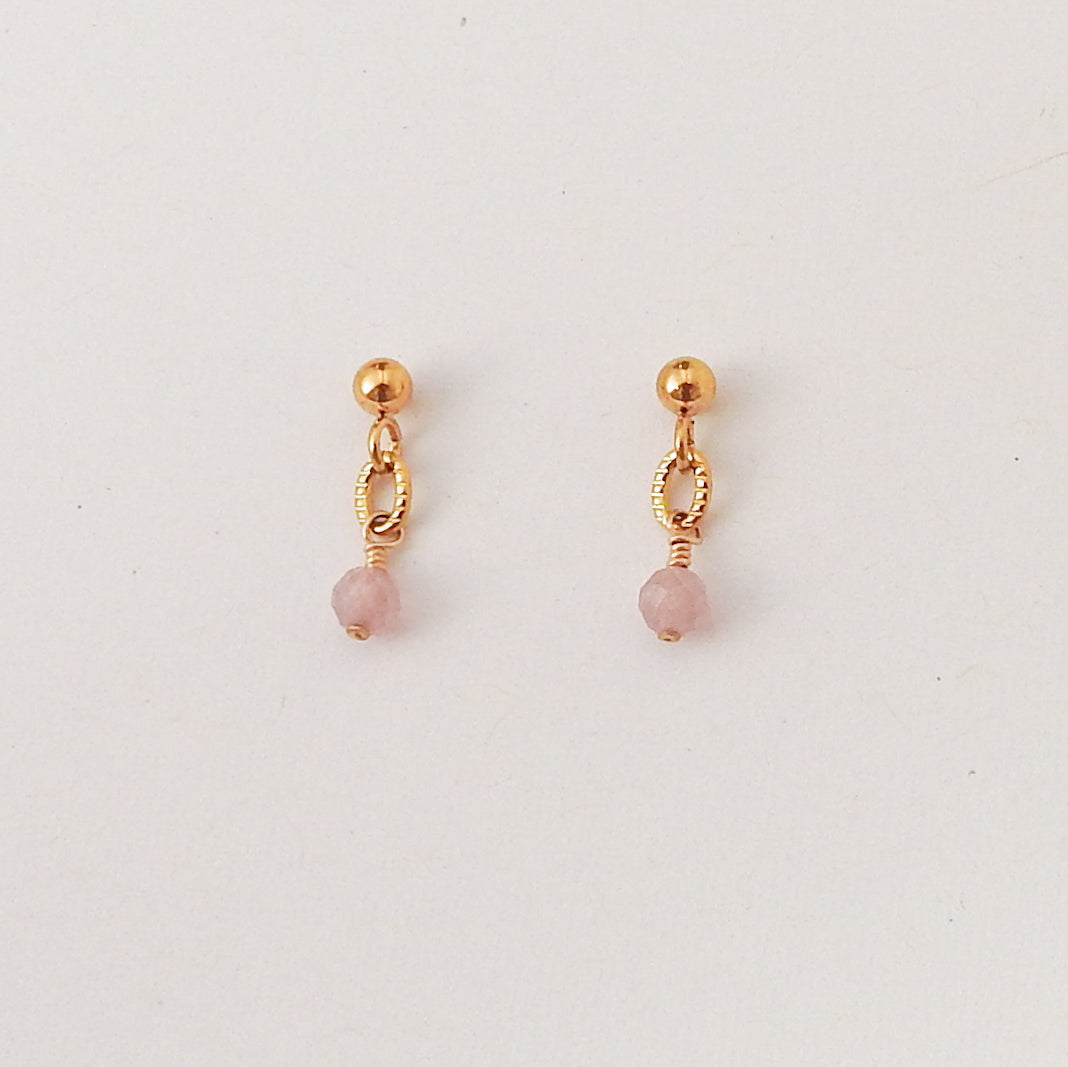 A pair of small gold dangle earrings with a faceted pink bead hanging from a textured oval link, displayed on a clean white background.