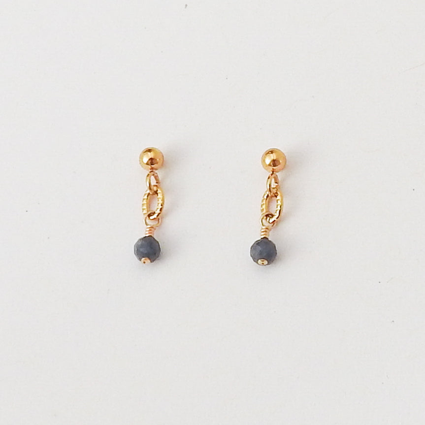 A pair of gold dangle earrings with a small gold ball, a twisted gold chain link, and a faceted dark gray bead dangling at the bottom, shown on a plain white background.