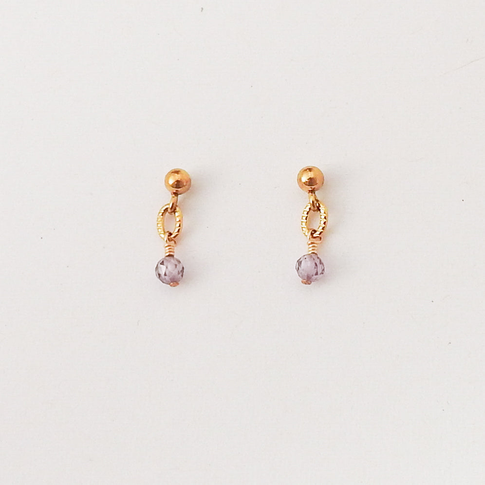 A pair of gold-toned dangle earrings with a small textured oval charm and a faceted light purple crystal drop, shown on a plain white background.