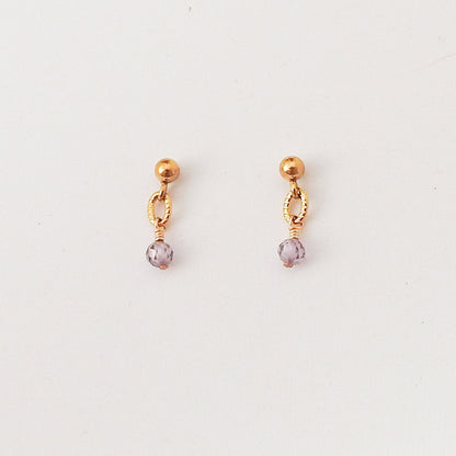 A pair of gold-toned dangle earrings with a small textured oval charm and a faceted light purple crystal drop, shown on a plain white background.