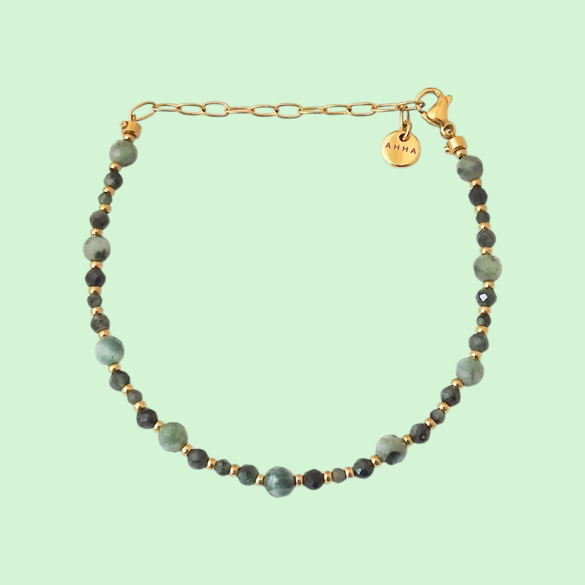 A beaded bracelet with light green and dark green faceted beads separated by small gold spacer beads, featuring a gold chain extender and a round gold charm engraved with AHHA