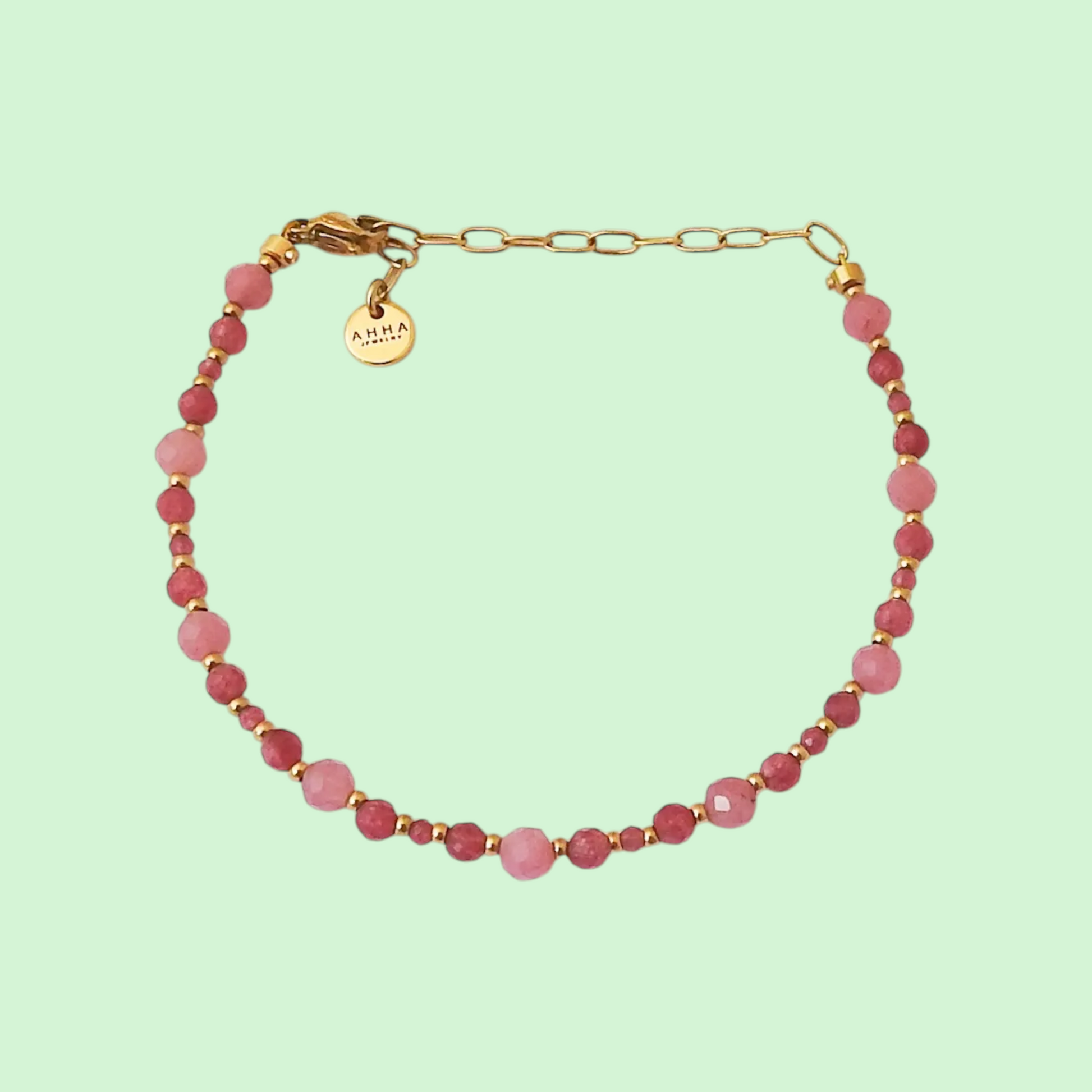 A delicate beaded bracelet with faceted pink beads and gold spacer beads, featuring a gold chain extender and a round gold tag engraved with 'AMIA' on a solid pale green background.