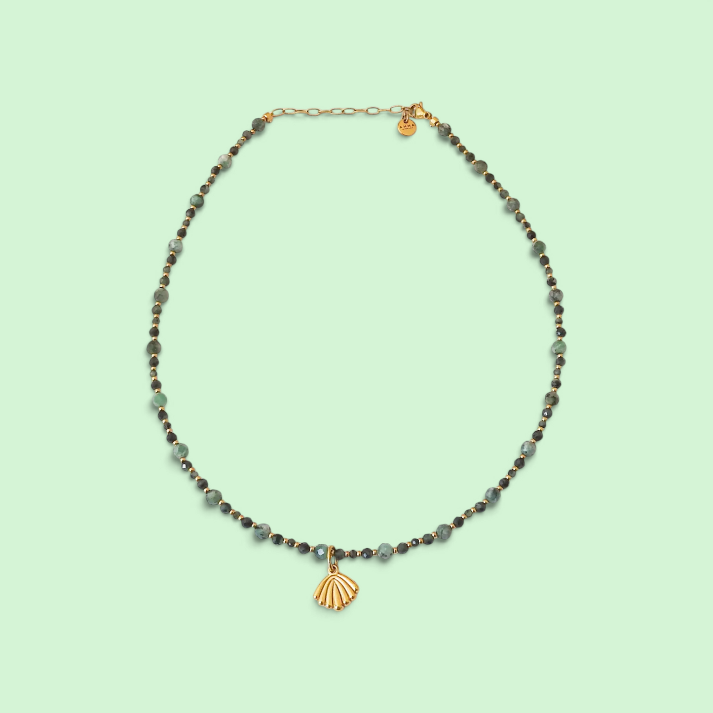 A delicate beaded necklace with gray and dark green faceted beads, a small gold heart charm, and a gold shell pendant, displayed on a solid pale mint green background.
