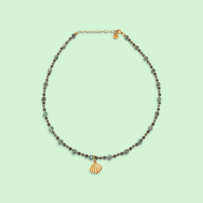 A delicate beaded necklace with gray and dark green faceted beads, a small gold heart charm, and a gold shell pendant, displayed on a solid pale mint green background.