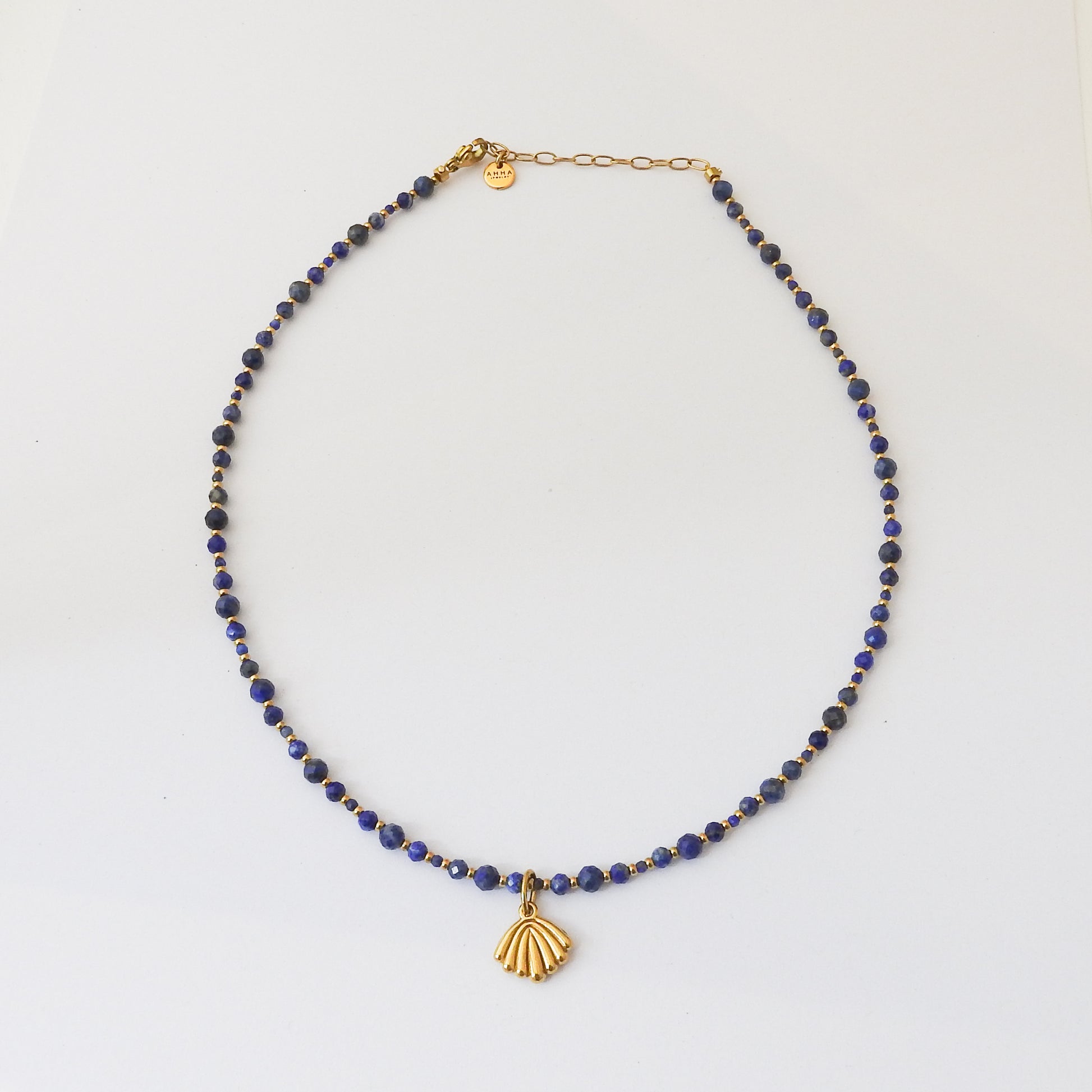 A beaded necklace featuring dark blue lapis lazuli stones and small gold spacer beads, with a gold shell-shaped pendant and gold chain clasp, displayed on a clean white background.