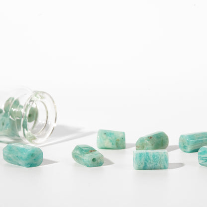 Turquoise rectangular gemstone beads with rounded ends, some lying on a white surface and one inside a clear glass jar, photographed on a clean white background.