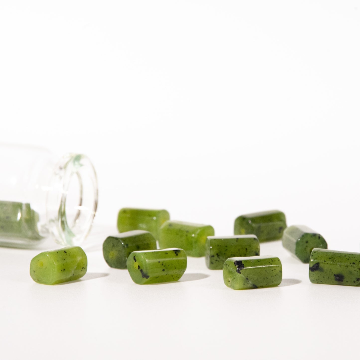 Green rectangular gemstone beads with dark flecks, shown spilling from a clear glass jar onto a white surface against a bright white background.